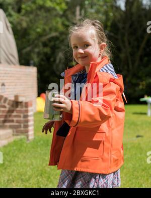 4 anni ragazza piantando e giardinaggio, piantando fuori piante di cetriolo, portando il cappotto di pioggia arancione/viola, giorno di sole, aiuti pre-schooler in giardino Foto Stock