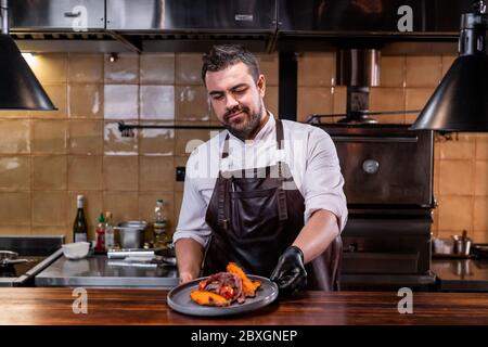 Chef con bearded in grembiule di pelle che mette il piatto preparato sul banco del ristorante Foto Stock