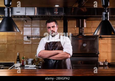 Ritratto di cuoco serio bearded in grembiule di cuoio in piedi con le braccia incrociate alla cucina del ristorante Foto Stock