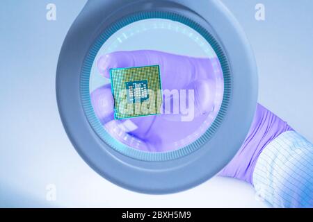 Visualizzazione in prima persona. Una mano in un guanto blu tiene un chip del processore e lo esamina sotto una lente di ingrandimento. Foto Stock