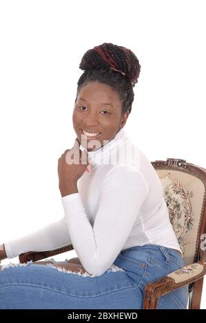 Una donna africana sorridente seduta su un top bianco e jeans in una vecchia poltrona, guardando nella macchina fotografica, isolato per sfondo bianco Foto Stock