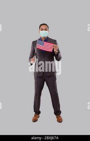 Uomo d'affari che tiene la bandiera americana che indossa la maschera medica. Uomo d'affari indiano in piedi a lunghezza intera con la bandiera degli Stati Uniti in mani Foto Stock