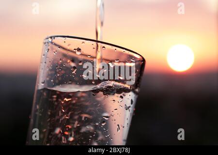 Versare acqua pulita nel bicchiere da bere sullo sfondo del tramonto. Concetto di salute e freschezza, sete, purificazione dell'acqua Foto Stock