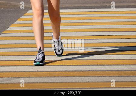 Ragazza sottile che attraversa la strada, gambe femminili in sneakers sulla traversata pedonale. Concetto di sicurezza stradale, donna in corsa nella città estiva Foto Stock