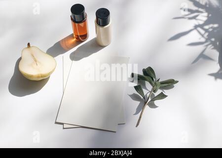 Stazioneria estiva scena ancora vita. Frutta di pera tagliata, olio cosmetico, bottiglie di crema e ramo di ulivo. Tavola bianca. Schede di carta bianche, moc inviti Foto Stock