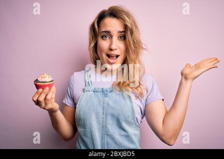 Giovane bella donna bionda eatimg cioccolato cupcake su isolato sfondo rosa molto felice ed eccitato, espressione vincitore che celebra la vittoria scre Foto Stock