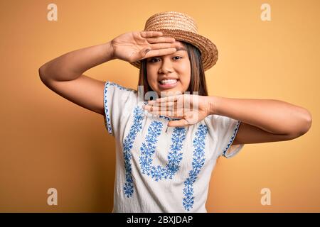 Giovane bella ragazza asiatica che indossa t-shirt casual e cappello in piedi su sfondo giallo sorridente allegro giocando a peek un boo con le mani che mostrano il volto. Foto Stock