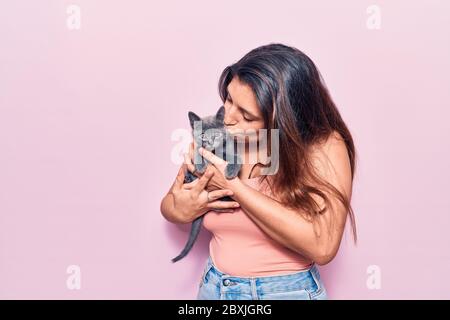 Giovane bella donna latina che puzzava felice. In piedi con sorriso sulla faccia che tiene e baciare adorabile gatto su isolato sfondo rosa Foto Stock