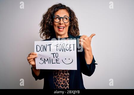 Donna di mezza età che tiene bandiera con non dimenticare il messaggio di sorriso su sfondo bianco che punta e mostra con il pollice fino al lato con il volto felice SM Foto Stock