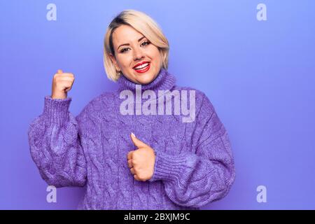 Bella donna bionda Plus taglia indossa un pullover casual a turleneck su sfondo viola che punta alla schiena dietro con mano e pollici in su, sorridente Foto Stock