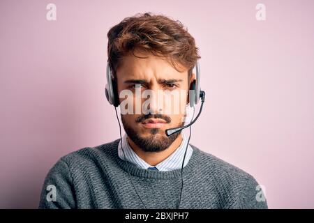Giovane agente del call center uomo con barba che indossa cuffie su uno sfondo rosa isolato, scettico e nervoso, disapprovando l'espressione sul viso con croce Foto Stock