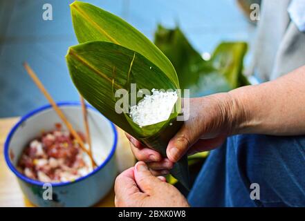 Foglia di tenuta a mano riempita di riso appiccicoso per fare Zong zi, gnocchi di riso cinese. Otturazioni di maiale sfocate sullo sfondo. Foto Stock