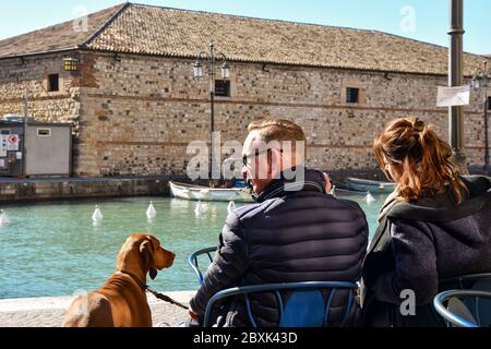 Una coppia con il cane in un caffè all'aperto che si affaccia sul canale, con la Casa di personalizzazione veneziana sullo sfondo, Lazise, Verona, Veneto, Italia Foto Stock