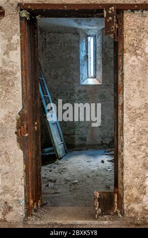 Una vecchia porta di legno con vernice che si appoggia contro il muro di una cella di prigione. La luce dalla finestra proietta interessanti ombre sulla parete. Foto Stock