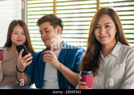 Una donna asiatica che tiene la tazza di caffè di carta con due amici che guardano lo stesso telefono cellulare in background Foto Stock
