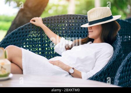 Immagine ritratto di una bella donna asiatica con cappello sdraiato su una panchina nel giardino con cocco sul tavolo Foto Stock