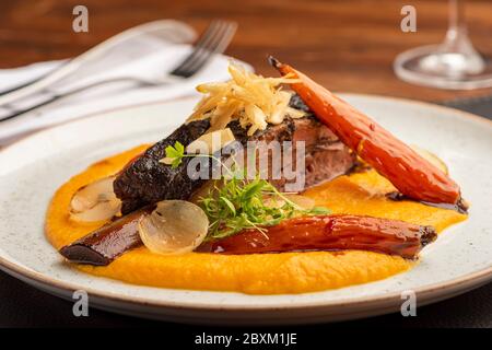 costolette guarnite con verdure su purè di patate su un piatto bianco con fondo sfocato con bicchiere di vino a forchetta Foto Stock