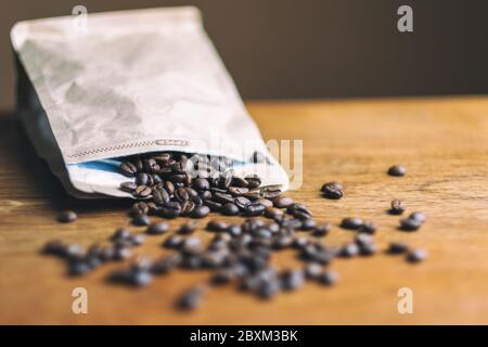 Il caffè in grani fuoriesce da un sacchetto di carta su un tavolo di legno Foto Stock