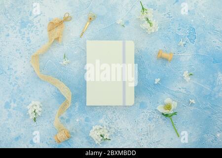 Composizione estiva di fiori con diario bianco, nappina e forbici dorate su sfondo blu. Disposizione piatta, vista dall'alto Foto Stock