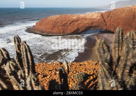 Vista del famoso arco sulla spiaggia di Legzira in Marocco. Cactus in primo piano. Regione Sidi Ifni. Popolare punto di riferimento turistico della costa marocchina. Foto Stock