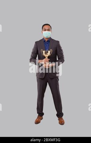 Businessman Holding Trophy indossare maschera medica. Uomo d'affari indiano in piedi a lunghezza intera con Trofeo in mani Foto Stock