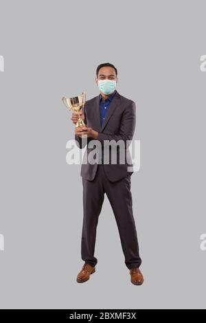 Businessman Holding Trophy indossare maschera medica. Uomo d'affari indiano in piedi a lunghezza intera con Trofeo in mani Foto Stock