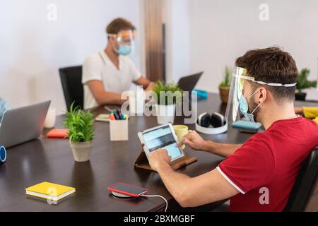 I giovani che lavorano all'interno di un ufficio creativo di codorking mentre indossano una maschera protettiva per la prevenzione della diffusione del coronavirus - Social Distancing, TECH A. Foto Stock