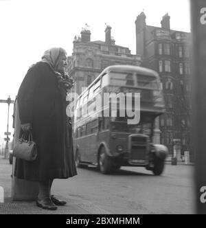 Londra negli anni '50. Vecchia donna con scialle a Londra 1952 in piedi per strada e un autobus a due piani passa. Foto Stock