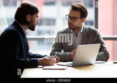 Due uomini d'affari sicuri seduti alla scrivania che parlano di risolvere le questioni comuni Foto Stock