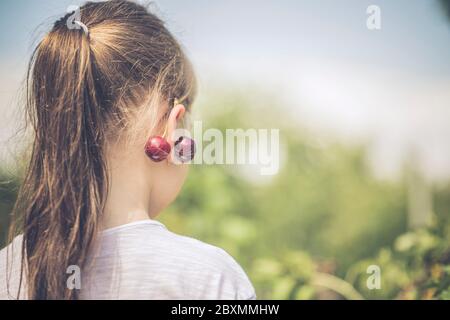 Bella bambina ha avuto una ciliegia fresca sull'orecchio. Adorabile bambina con una ciliegia sull'orecchio. Fare gioielli da frutta. Foto Stock