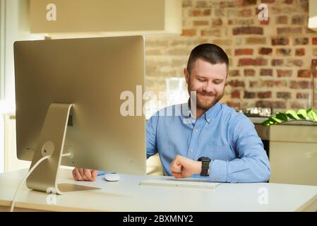 Un uomo felice con una barba che guarda il suo orologio da polso durante un video briefing online con i suoi colleghi a casa. Un ragazzo con un sorriso che lavora a distanza Foto Stock