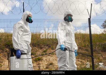Ritratto di due lavoratori che indossano dispositivi di protezione e spruzzano sostanze chimiche all'aperto in piedi da recinzione di filo spinato durante la disinfezione o la pulizia in mite Foto Stock
