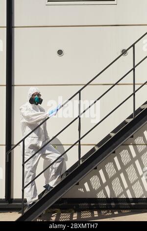 Verticale verticale verticale a lunghezza intera del lavoratore che indossa un muta in piedi su scale all'aperto alla luce del sole durante la disinfezione o la pulizia, spazio per la copia Foto Stock