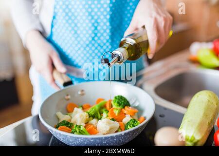 Donna che versa olio d'oliva sulle verdure in padella a casa. Concetto di pasto sano. Foto Stock