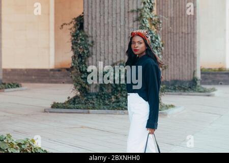 Bella giovane ragazza musulmana africana nera che indossa una bandana sulla strada della città moderna. Cammina in jeans bianchi con un sorriso felice sul viso. FAS Foto Stock
