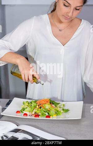Messa a fuoco bella giovane donna aggiungendo olio di oliva ad un'insalata appena fatta su un piatto bianco alla cucina su uno sfondo fuori fuoco. Cucinare a casa Foto Stock