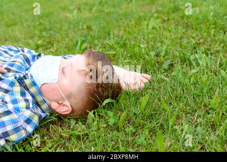 Ritratto di un ragazzo con maschera chirurgica adagiata sull'erba del parco. Primo piano di un ragazzino in una camicia a plaid che si rilassa sull'erba. Foto Stock