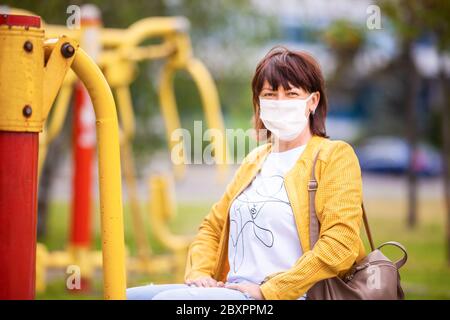 Donna di mezza età non identificata in abiti casual cammina attraverso il parco primaverile in una maschera medica bianca protettiva. Concetto di protezione del Coronovirus Foto Stock