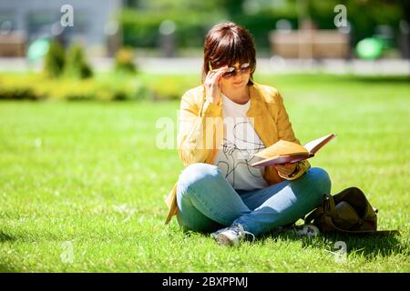 Donna di mezza età positiva in occhiali da sole si siede su un campo in un parco e legge un libro in una calda giornata estiva soleggiata. Concetto di hobby e attività all'aperto Foto Stock