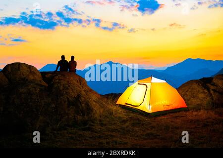 Giovane coppia seduto insieme sulla cima della roccia vicino tenda illuminata escursionistica e godendo la bella vista serale delle montagne sotto il cielo di tramonto Foto Stock