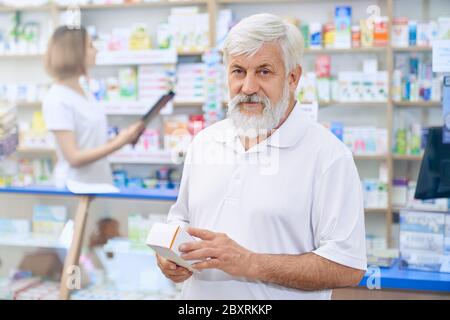 Ritratto di uomo anziano che sceglie farmaci in farmacia, guardando la macchina fotografica. Cliente senior con bearded in camicia bianca con scatola medica. Farmacista femminile in piedi al banco sullo sfondo. Foto Stock