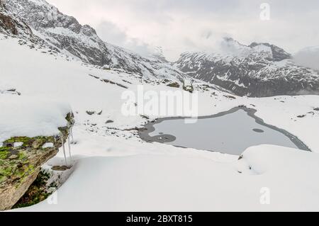 Vista su un laghetto innevato sulle montagne con un icicolo che indossa rocce mussose in primo piano. Foto Stock