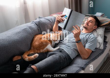 Gatto bianco e marrone si addormenta sul suo proprietario che legge sdraiato su un divano sotto la finestra. Foto Stock