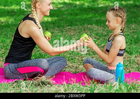 Madre e sua figlia in abbigliamento sportivo che tiene una mela verde e si siede sul tappeto in parco. Foto Stock