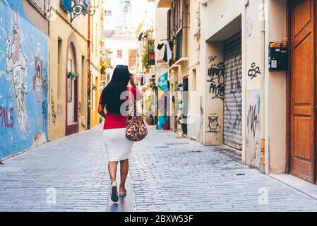 Brunette ragazza latina con borsa colorata che cammina sulla schiena lungo la stretta strada della città con pavimento in ciottoli. Concetto di turismo urbano Foto Stock