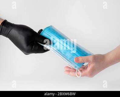 Una mano in una mano in un guanto nero in nitrile dà una maschera medica per proteggere il viso di un'altra persona.un guanto nero in nitrile dà una maschera medica per pro Foto Stock