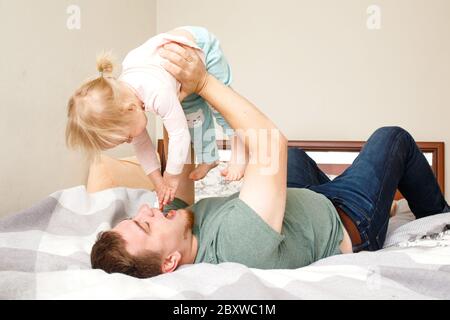 Giovane padre sdraiato su un letto e tenendo la sua figlia bambino capretto sulle mani, ridendo e divertendosi, concetto di festa del padre`s giorno, routine mattutina a casa Foto Stock