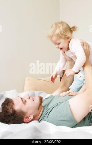 Giovane bel padre che si stette su un letto e tiene la sua bambina carina bambino sulle mani, ridendo e divertendosi, padre`s giorno concetto di vacanza, mattina r Foto Stock