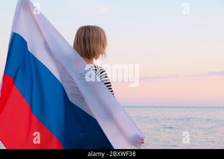 Indietro vista bionda donna che sventola bandiera nazionale Russia all'aperto tramonto mare in estate - bandiera russa, paese, patriottismo, Russia giorno e 12 giugno. Foto Stock