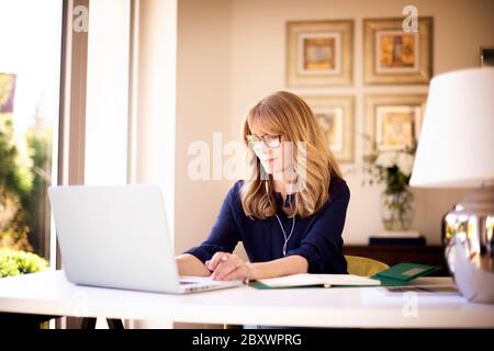 Ripresa di una donna d'affari di mezza età con auricolare mentre si siede dietro il suo laptop e si sta discutendo e incontrando online in videochiamata. Donna d'affari Foto Stock